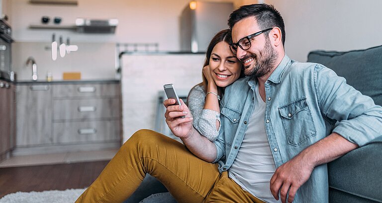 Paar sitzt im Wohnzimmer und schaut gemeinsam aufs Handy
